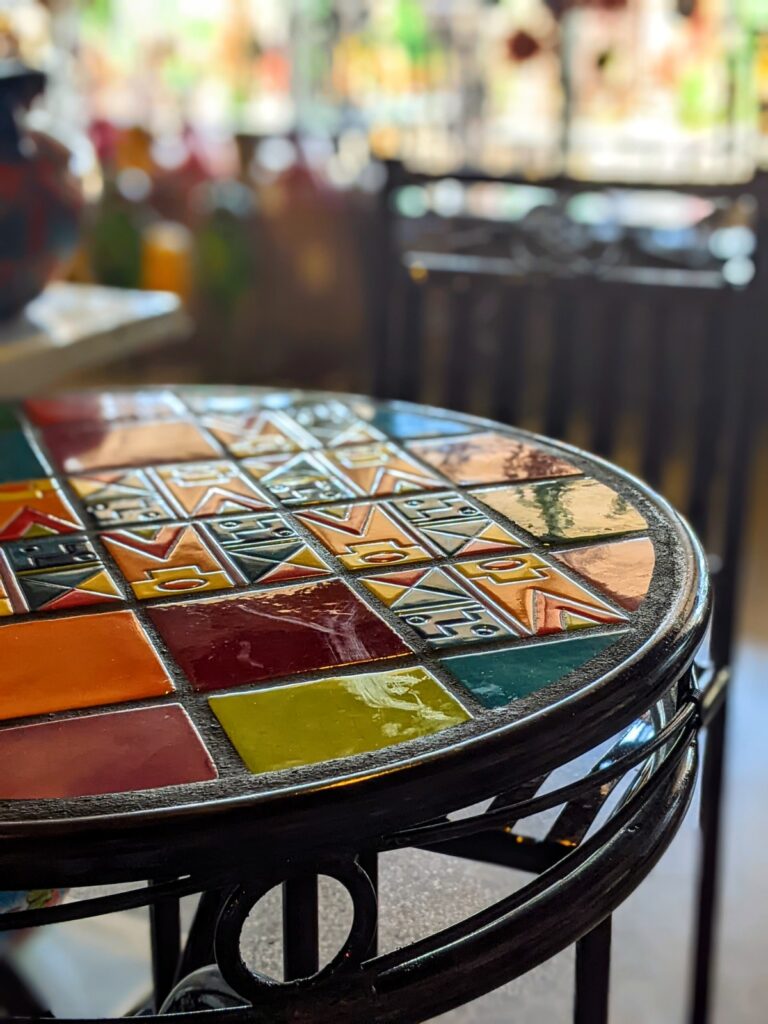 A round metal table with colorful tiles laid into it and a metal chair next to it.